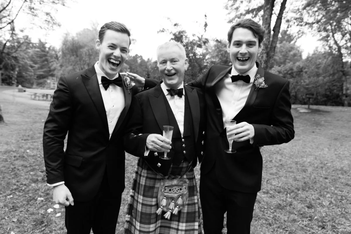 Three men in kilts and tuxedos smile for the camera.
