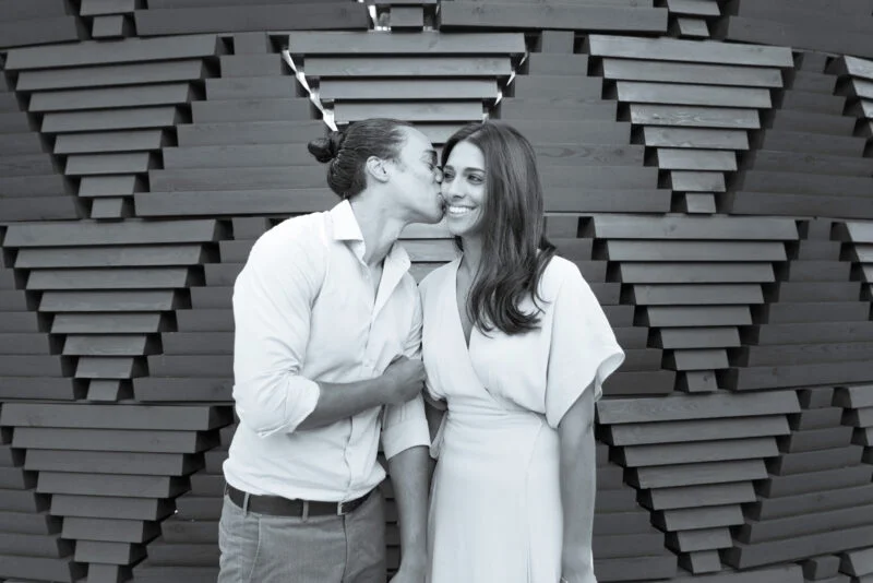 A man and woman kissing in front of a wooden structure.