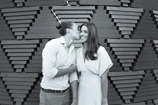 A man and woman kissing in front of a wooden structure.