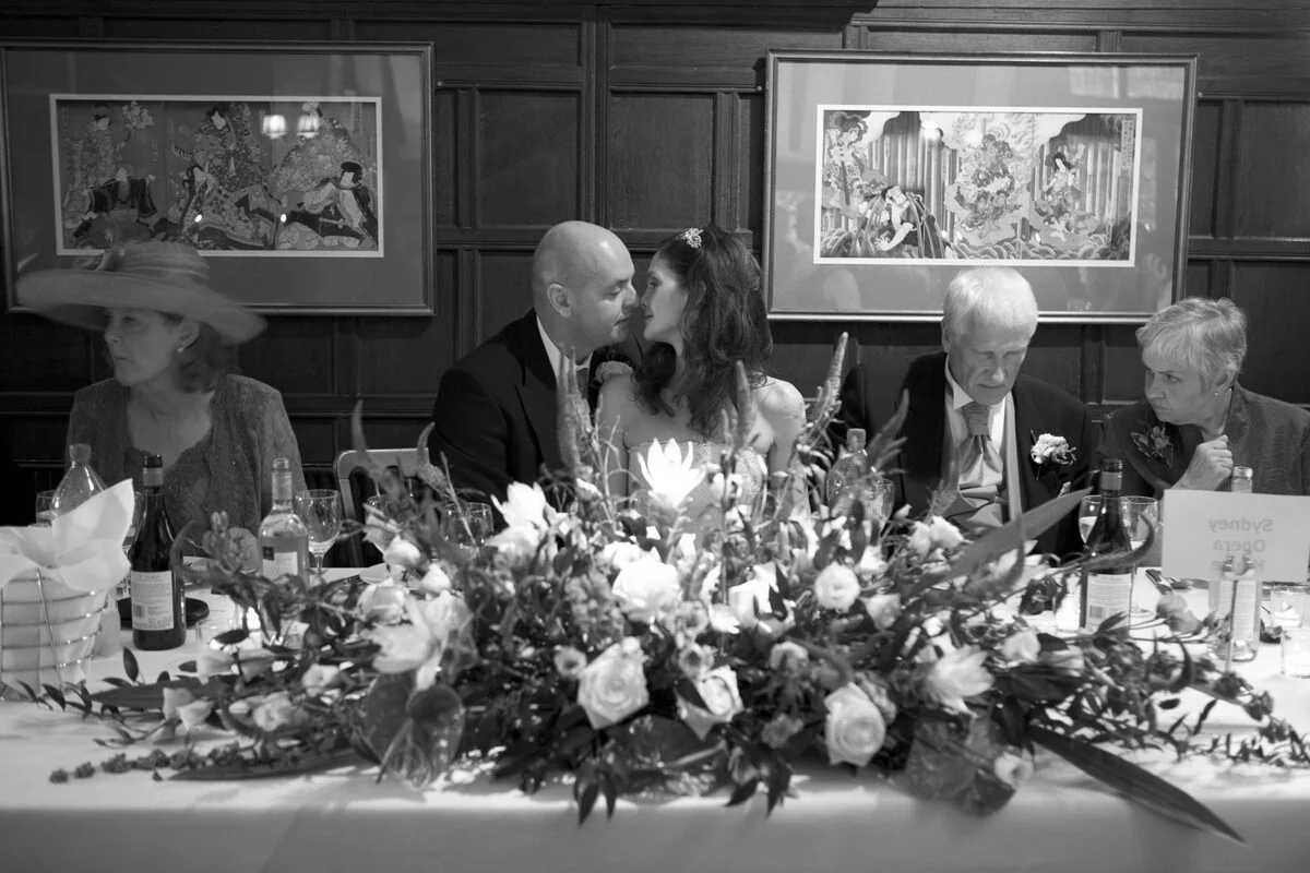 A black and white photo of a couple kissing at a table.