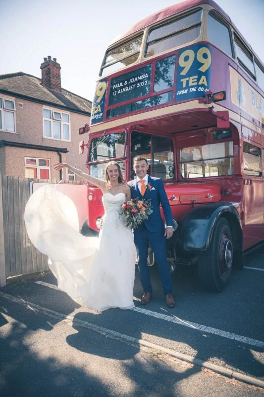 A bride and groom standing next to a double decker bus.
