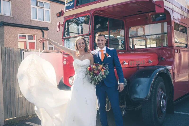 A bride and groom standing next to a double decker bus.