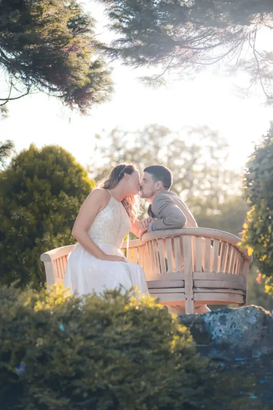 A bride and groom kiss on a bench in a garden.