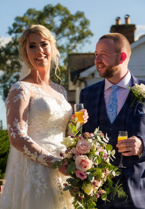 A bride and groom holding champagne glasses in front of a house.