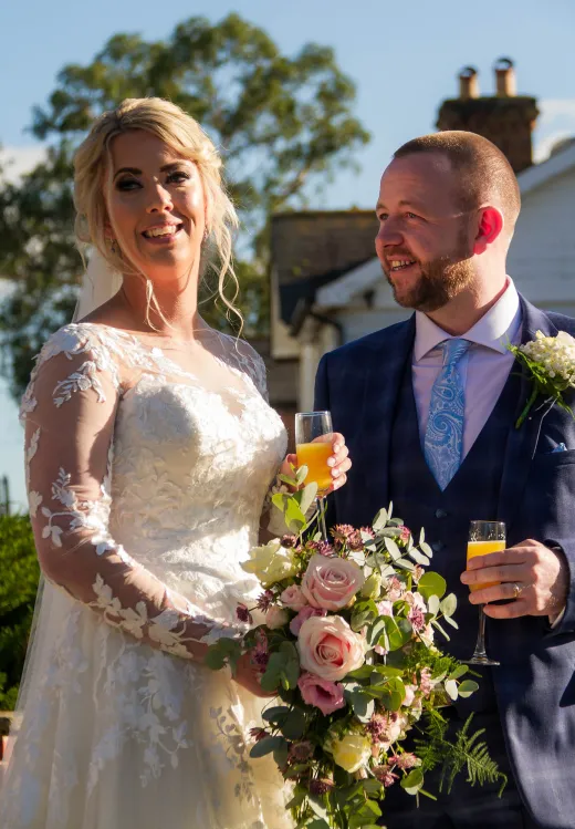 A bride and groom holding champagne glasses in front of a house.