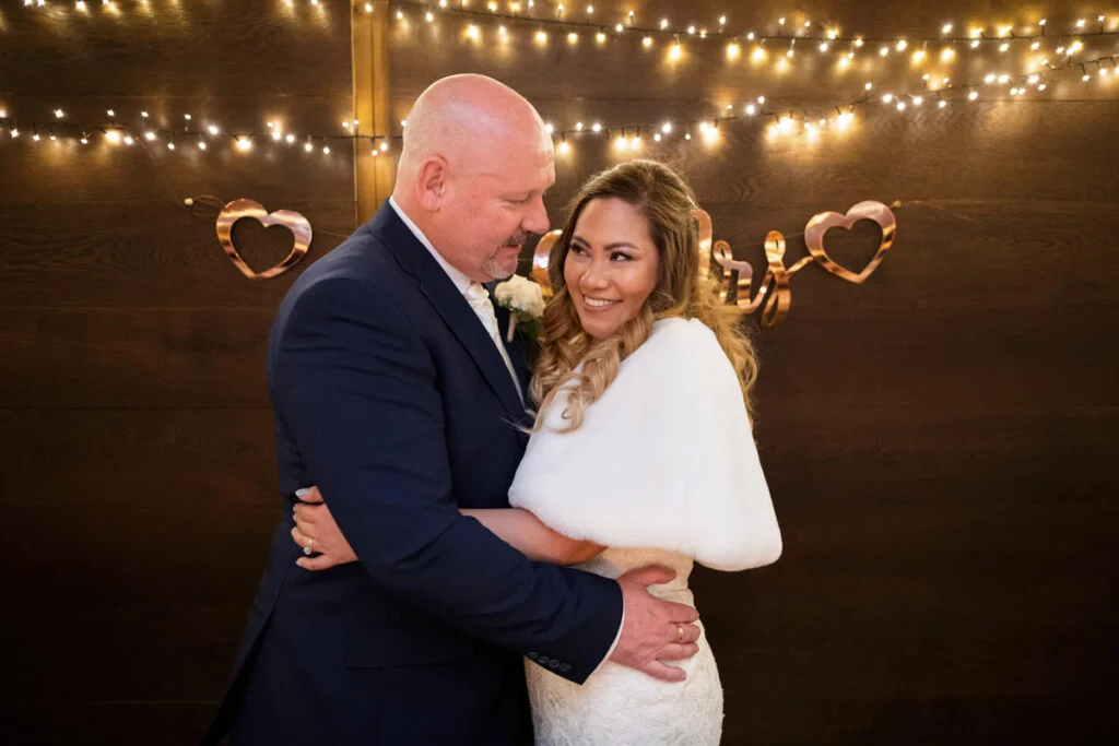A bride and groom hugging in front of a string of lights.