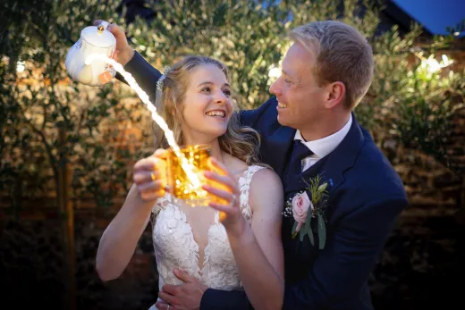A joyful bride and groom share a playful evening moment outdoors as glowing liquid is poured from a teapot into a mug.