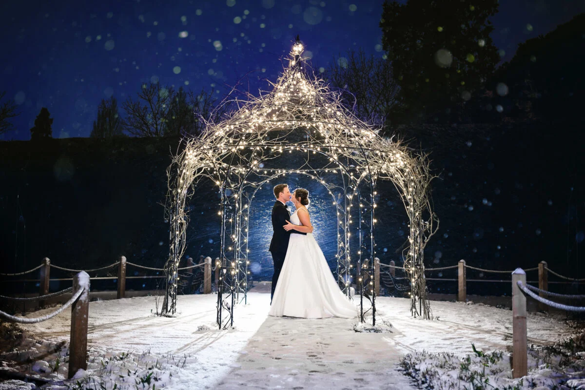 A bride and groom standing under a snow covered gazebo.
