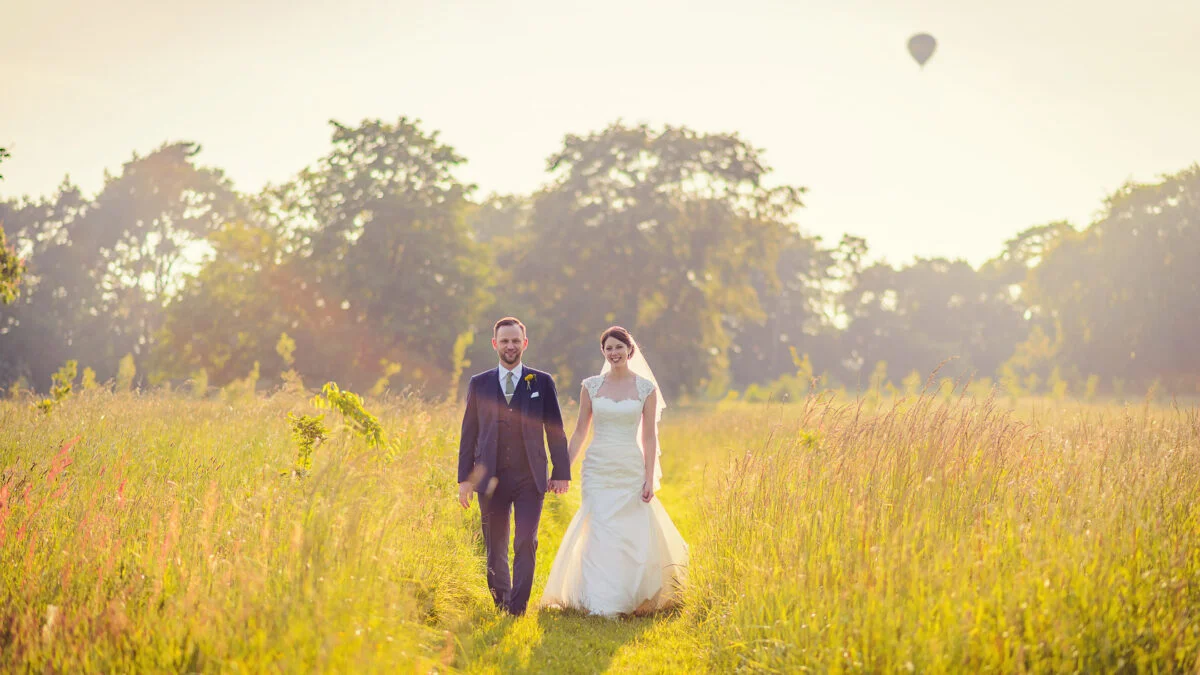 A bride and groom walking through a field with a hot air balloon.