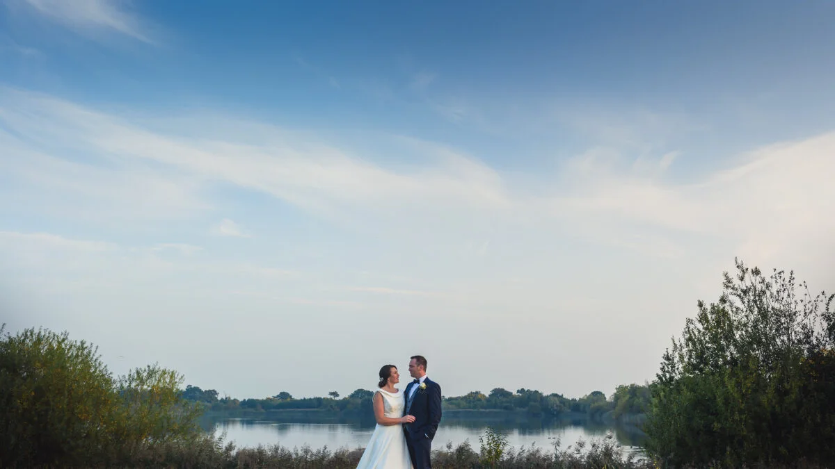A bride and groom standing in front of a lake.