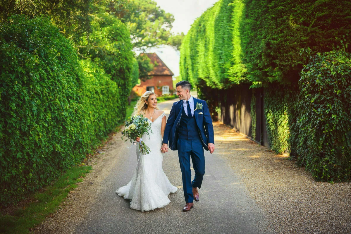 A bride and groom walking down a path surrounded by hedges.
