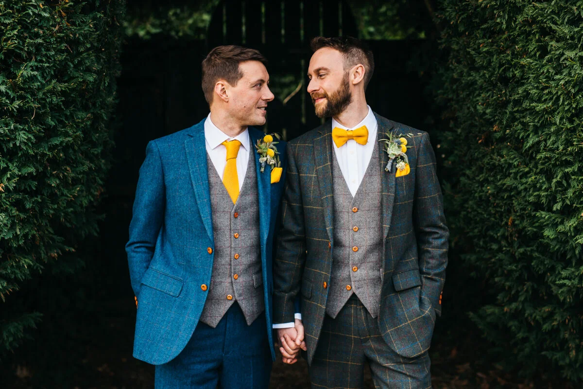 Two grooms wearing suits and bow ties in front of hedges.