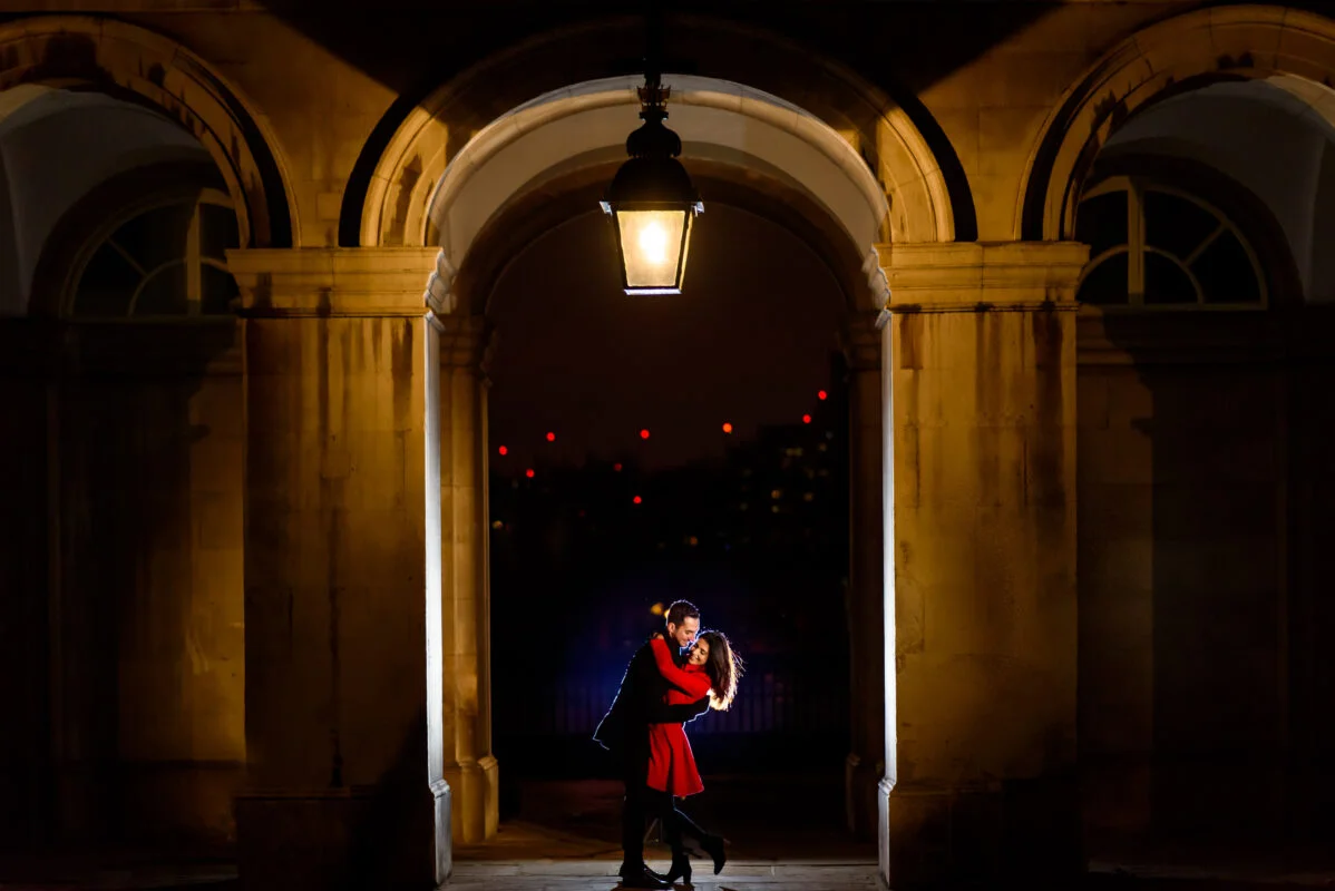 A couple kissing under an archway at night.