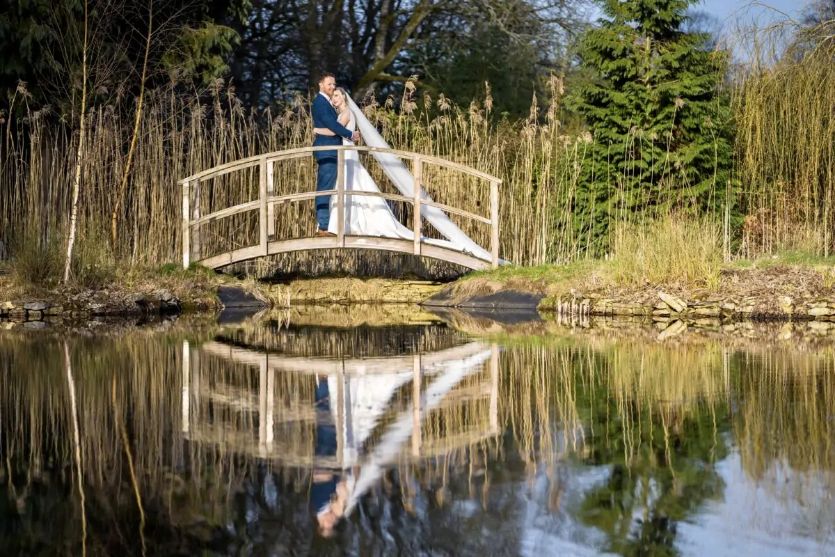 A bride and groom standing on a bridge over a pond.
