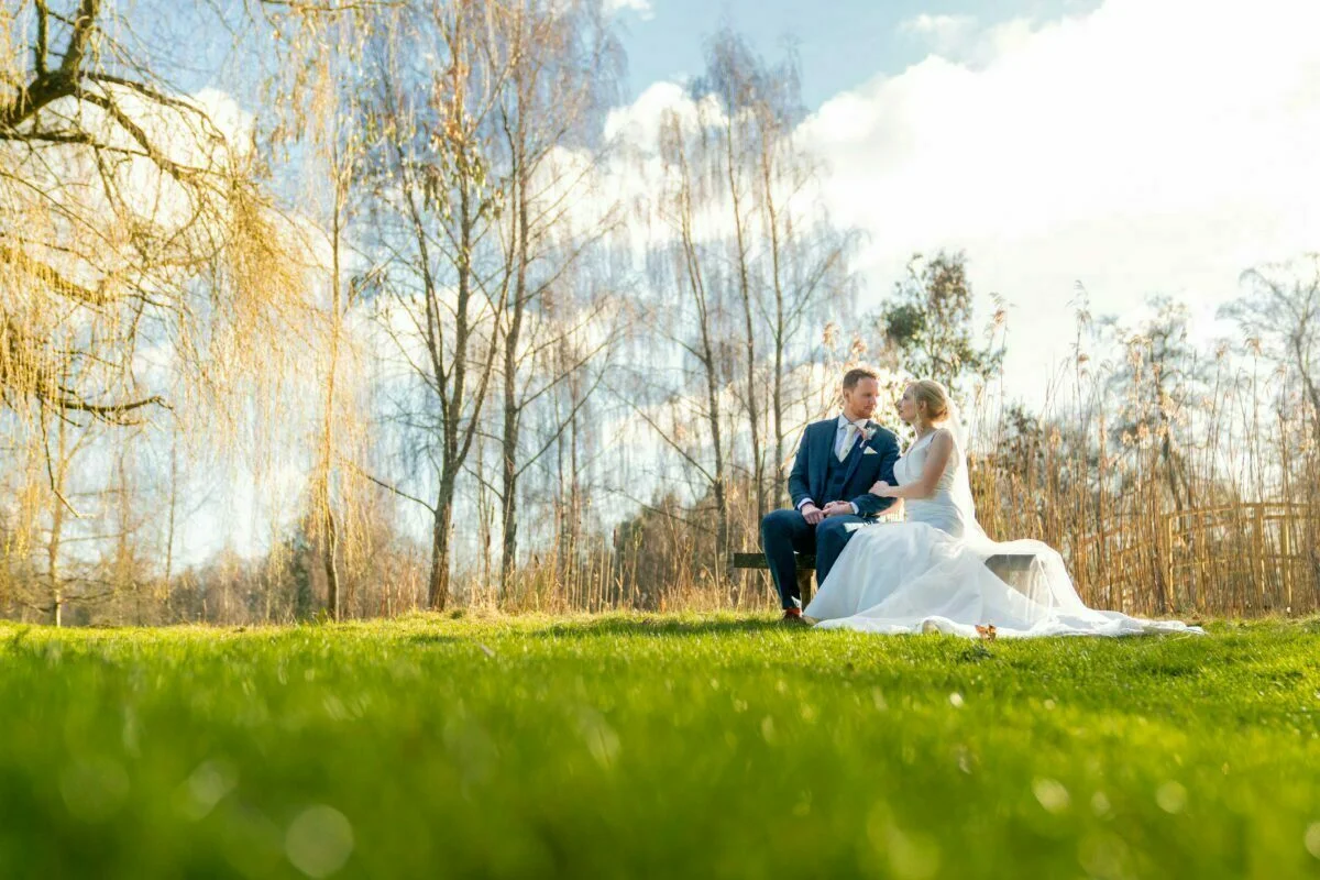 A bride and groom sitting on a bench in the grass.