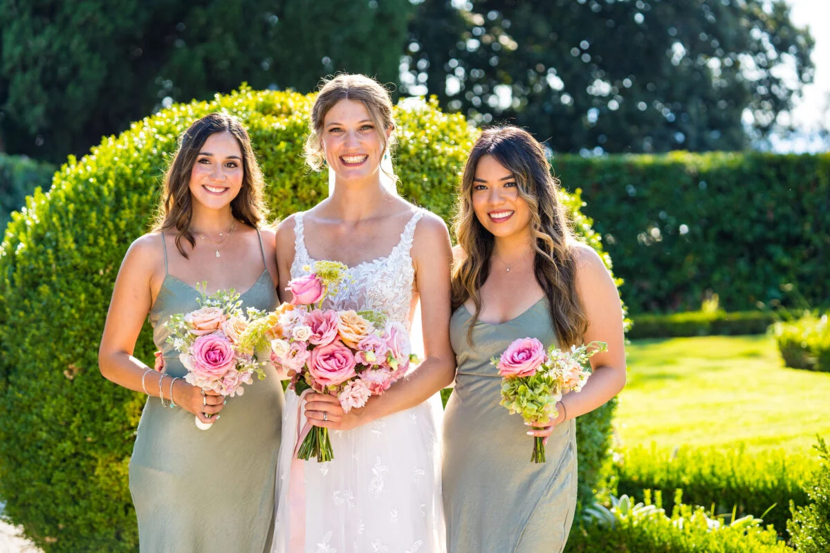 Three bridesmaids pose with their bouquets in front of a hedge.