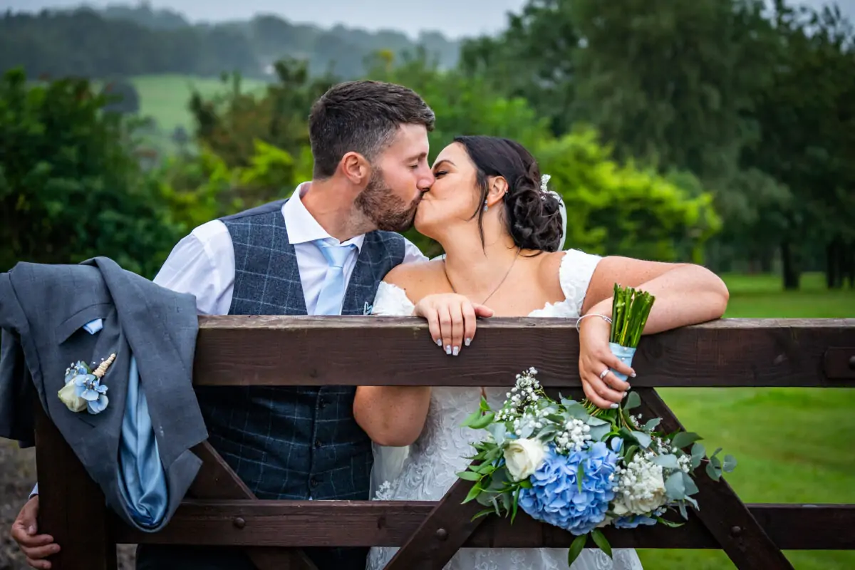 A bride and groom kissing on a wooden fence.