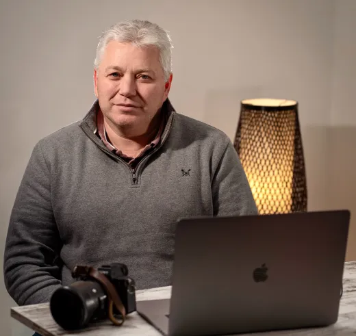 A mature man with grey hair wearing a grey jumper sits at a desk with a laptop and camera.