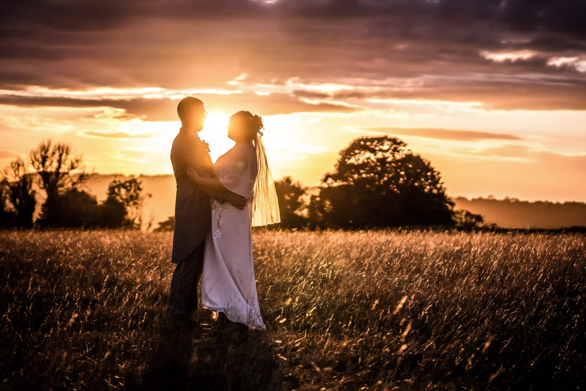 A bride and groom standing in a field at sunset.