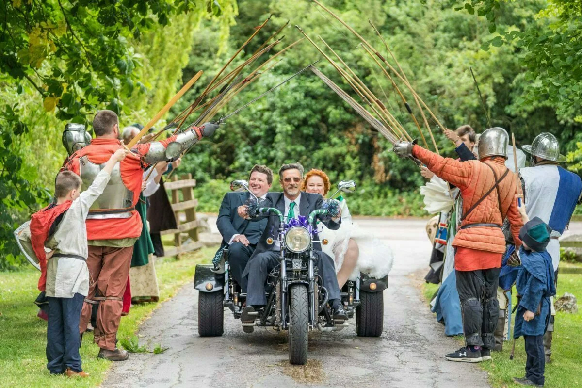 A bride and groom on a motorcycle with swords in their hands.