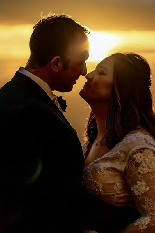 A bride and groom kissing in front of a sunset.