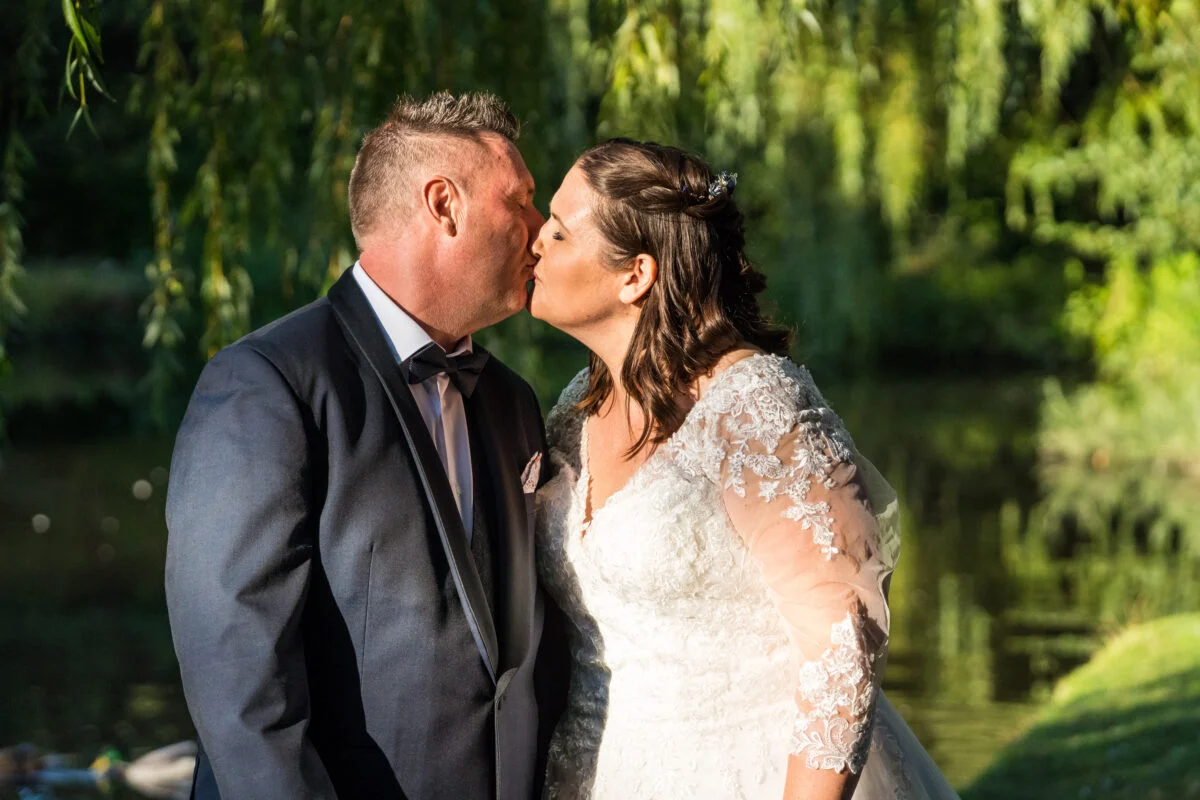 A bride and groom kiss in front of a willow tree.