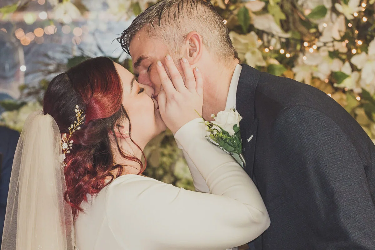 A bride and groom kissing in front of a floral backdrop.