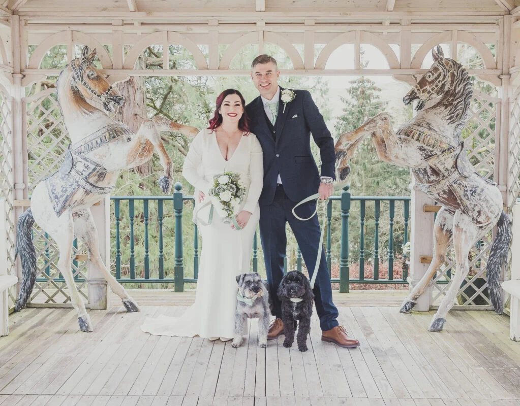 A bride and groom pose with their dog in front of a carousel.