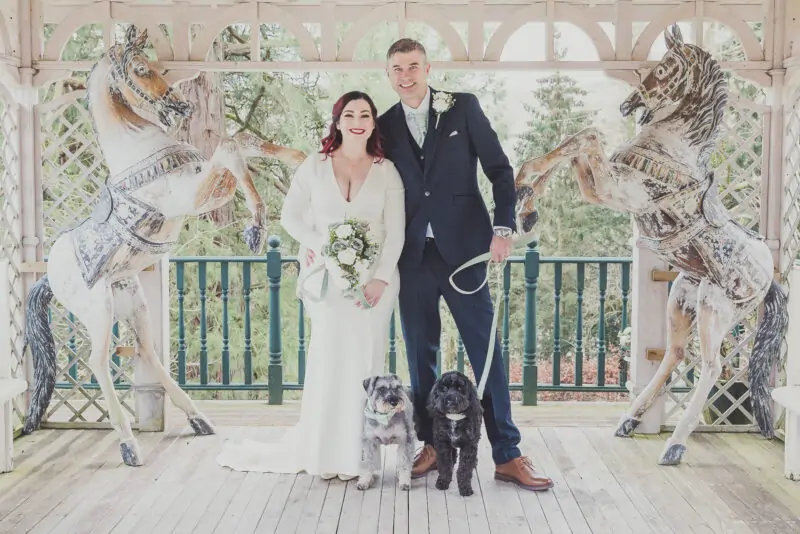 A bride and groom pose with their dog in front of a carousel.