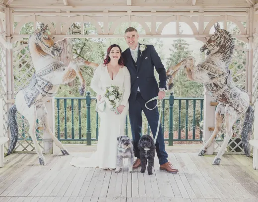 A bride and groom pose with their dog in front of a carousel.