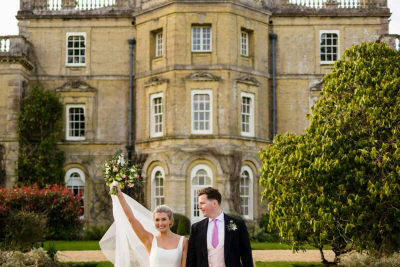 A bride and groom standing in front of a large mansion.
