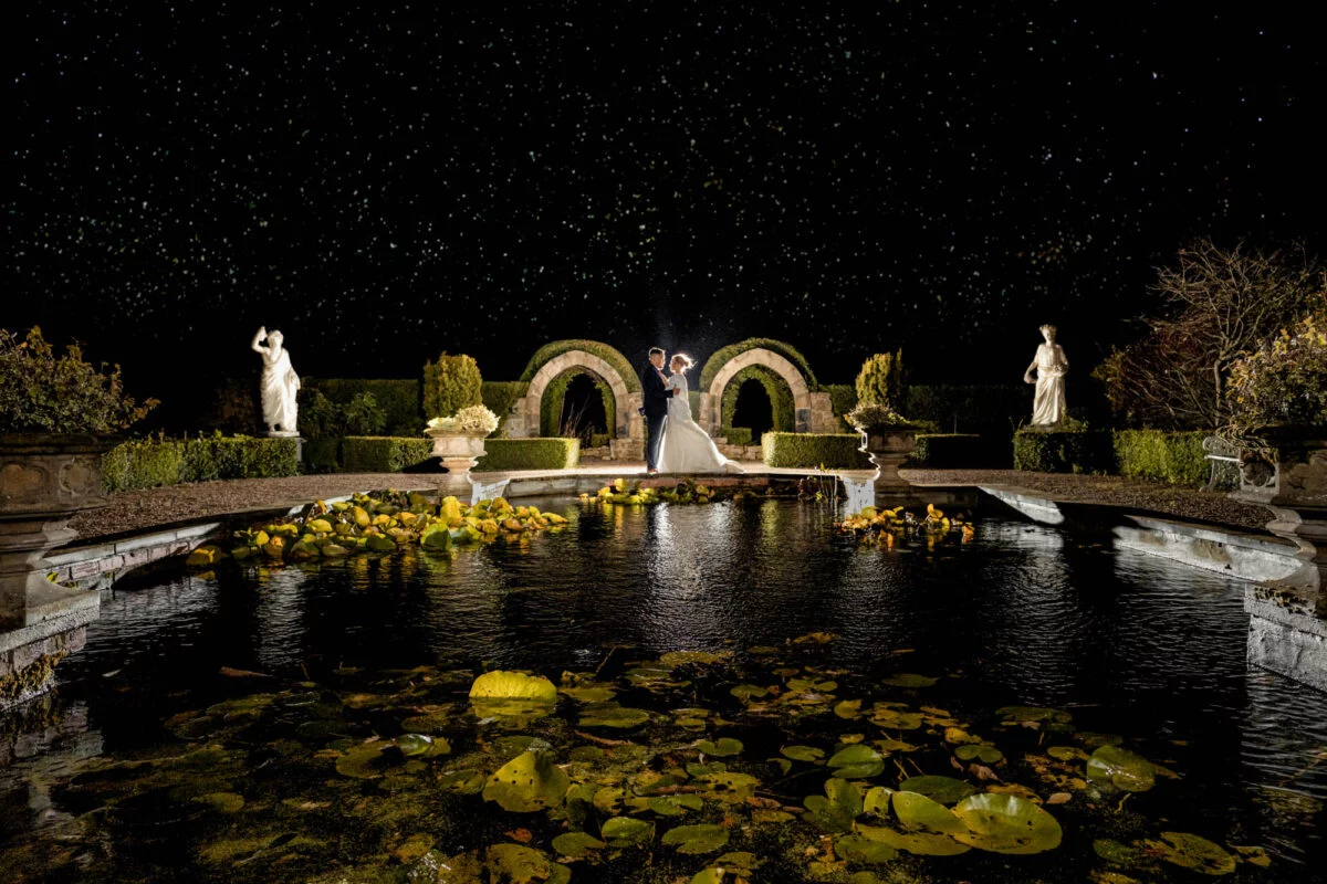 A bride and groom standing in front of a pond at night.