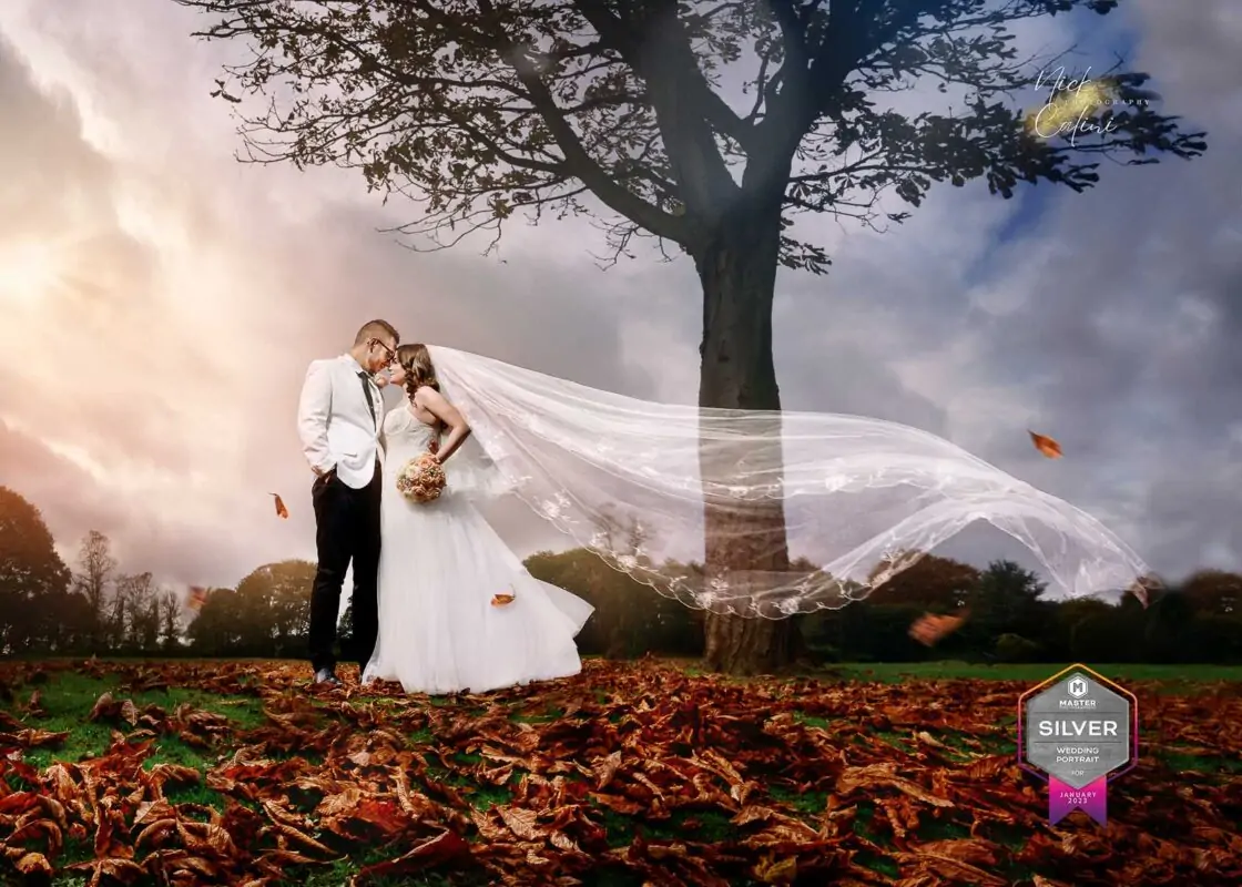 A bride and groom standing under a tree with a veil blowing in the wind.
