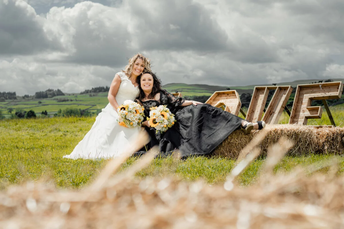 Two brides posing for a photo in a field with hay bales.