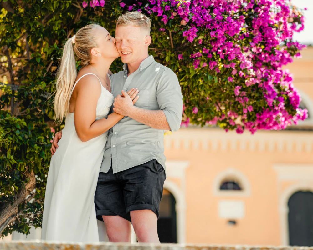 A couple kisses in front of a purple flowering tree.