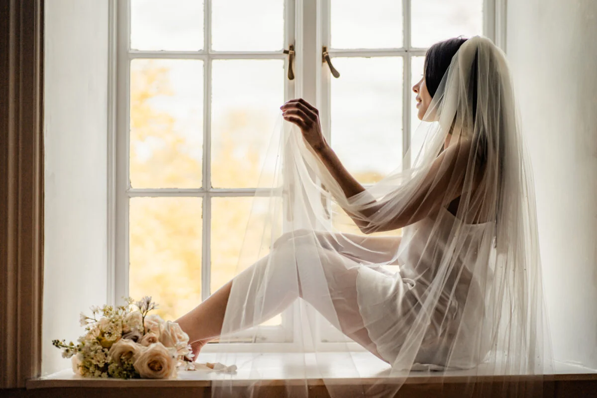A bride in a wedding dress sits on a window sill.