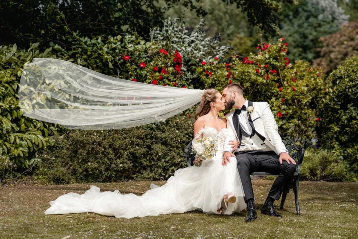 A bride and groom sitting on a bench with their veil blowing in the wind.
