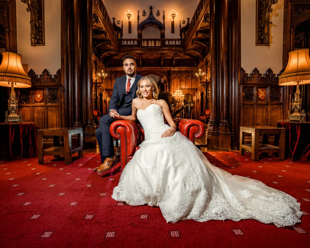 A bride and groom sitting on a red chair in an ornate room.