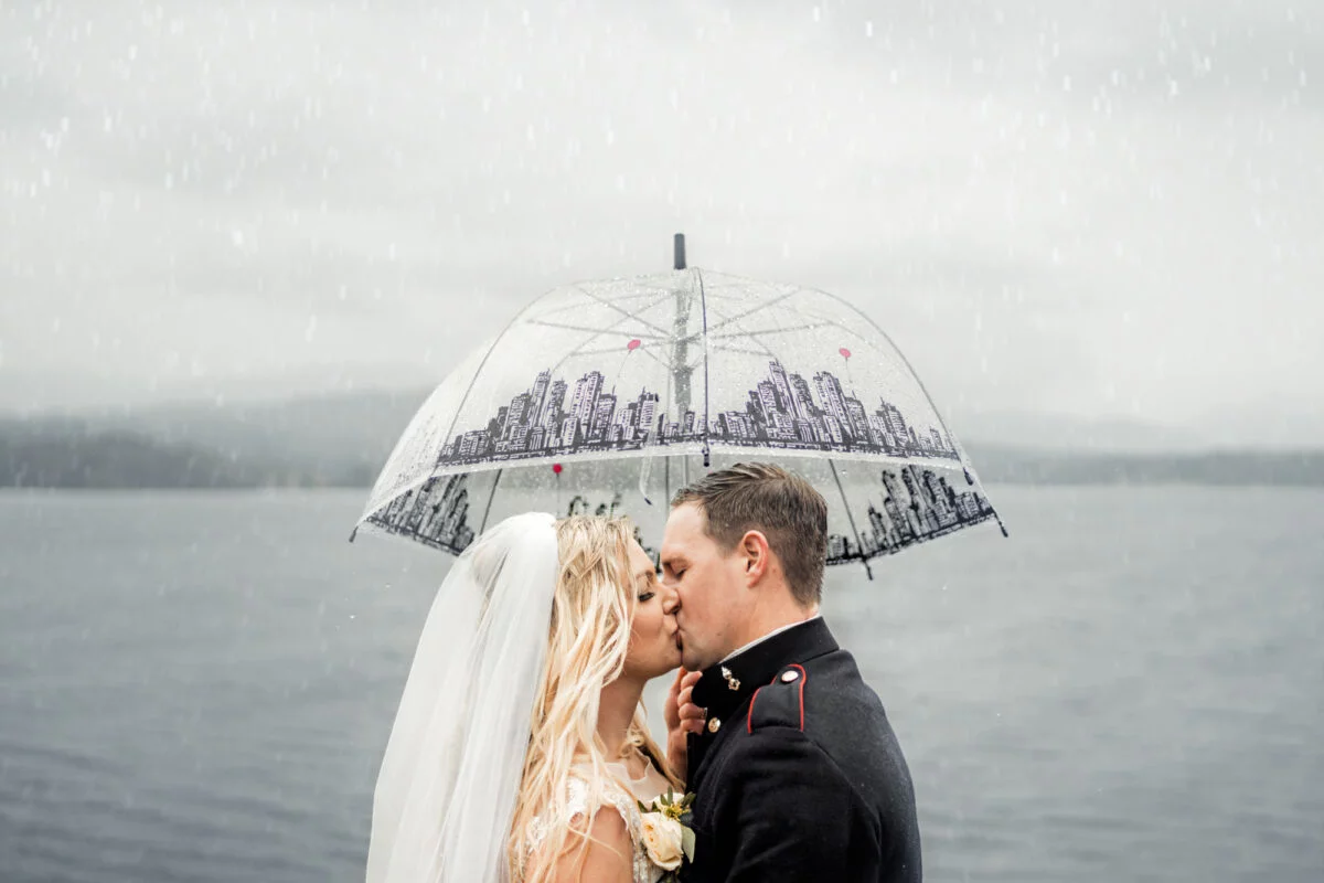 A bride and groom kiss under an umbrella in the rain.
