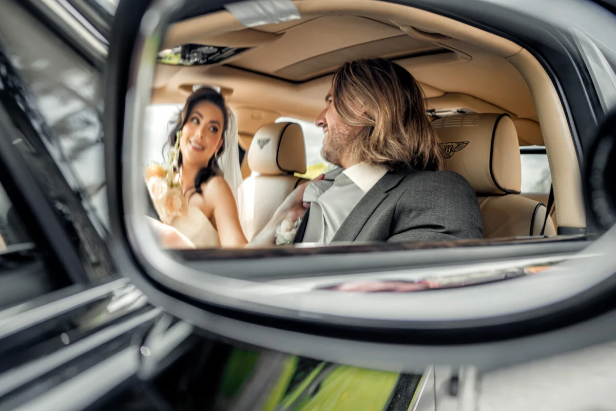 A bride and groom looking at each other in the rear view mirror of a car.
