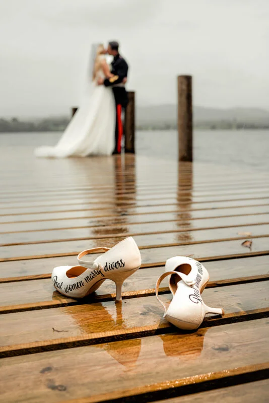 A bride and groom standing on a dock with their wedding shoes.