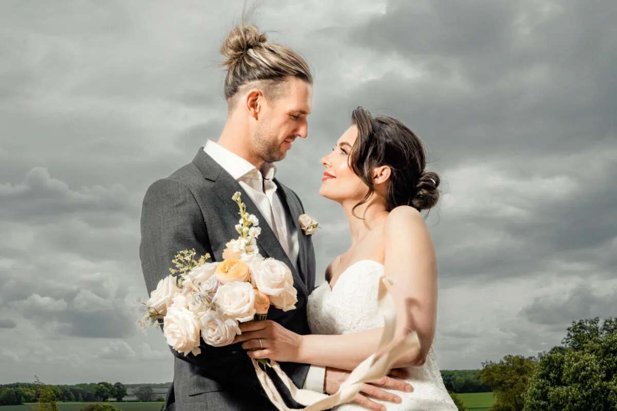 A bride and groom posing in front of a cloudy sky.