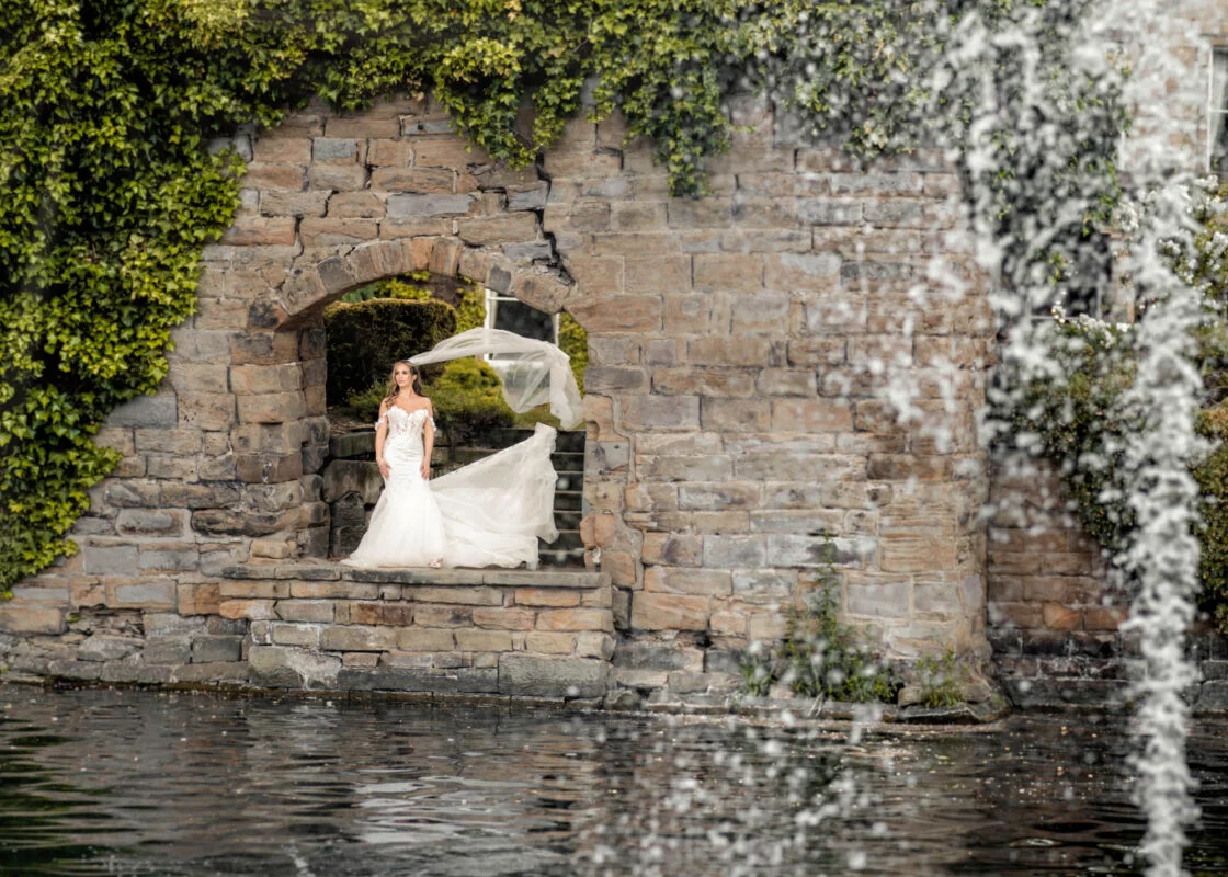 A bride is standing in front of a fountain in a stone wall.