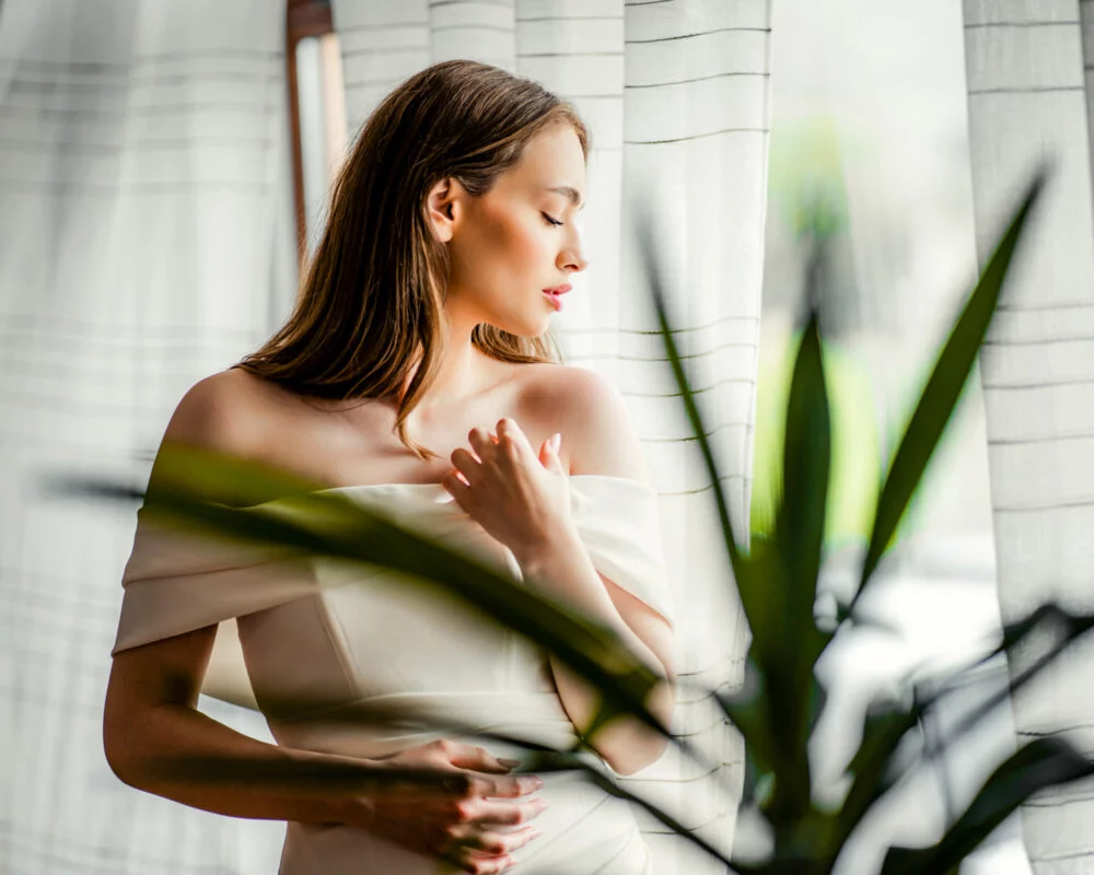 A beautiful woman in a white dress standing by a window.