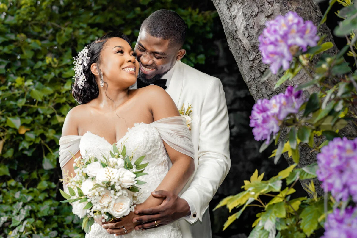 A bride and groom embrace in front of purple flowers.