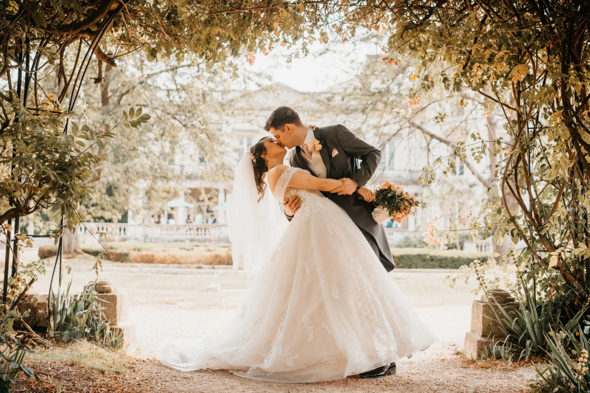 A fairytale bride and groom kissing under an archway in a garden captured through wedding photography. (listing)