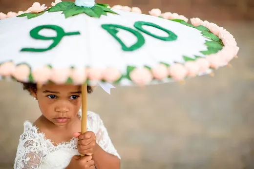 An adorable flower girl in a lace dress holds a decorated white parasol featuring green lettering and pink floral trim.