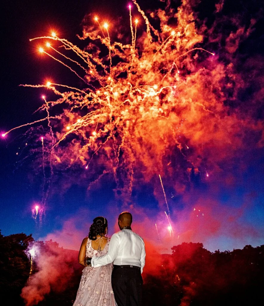 A wedding photographer duo capturing a bride and groom in front of a firework display. (listing)