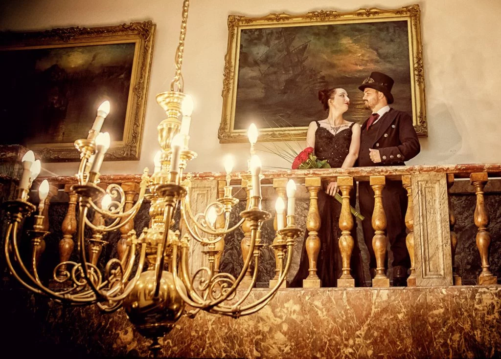 A couple standing on a balcony in front of an ornate chandelier.