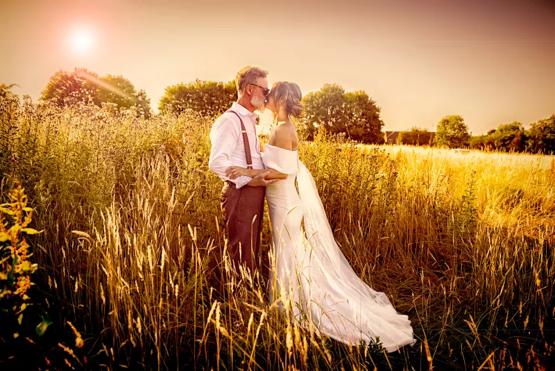 A wedding photographer duo capturing a bride and groom kissing in a field of tall grass. (listing)